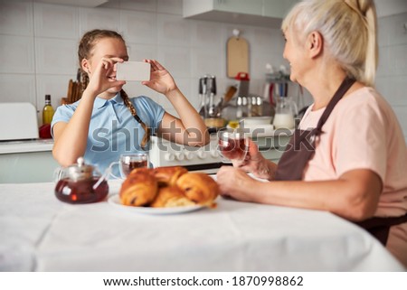 Lovely grey-haired lady posing with cup of tea while a girl taking her picture on smartphone