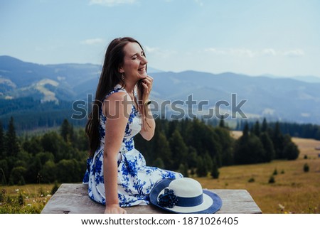 young woman in elegant white and blue dress sitting on bench in the mountains and smiling. Female relaxing in the top of Carpathians. travel to the mountains on a warm sunny summer's day