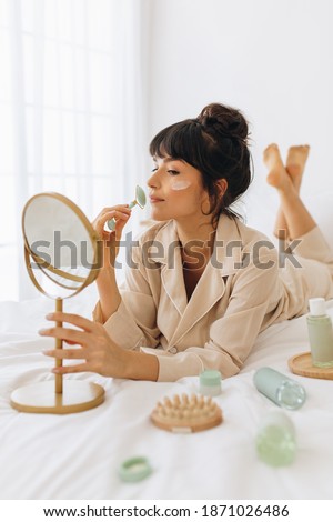 Woman using roller to massage face. Close up of woman applying face cream looking into a mirror lying on bed at home. Royalty-Free Stock Photo #1871026486