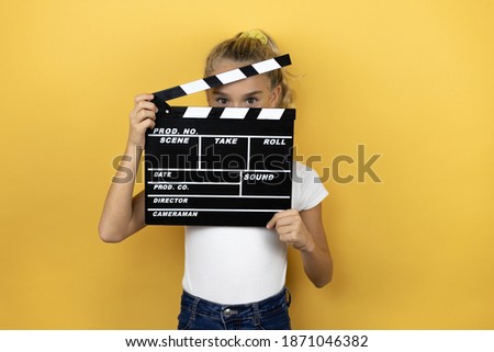 Young beautiful child girl standing over isolated yellow background Young beautiful child girl standing over isolated yellow background holding a clapperboard very happy
