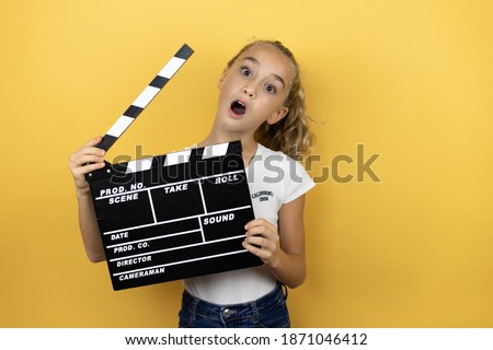 Young beautiful child girl standing over isolated yellow background holding a clapperboard very happy