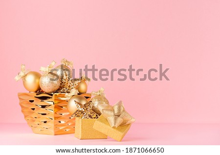 Luxury New Year gold shimmer decor with christmas stars, balls with ribbon in bowl, present on soft light pink background.