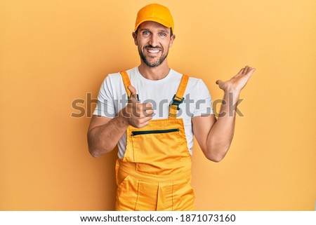 Young handsome man wearing handyman uniform over yellow background showing palm hand and doing ok gesture with thumbs up, smiling happy and cheerful 