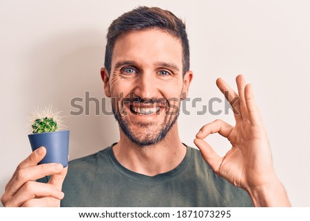 Young handsome man holding small cactus plant pot over isolated white background doing ok sign with fingers, smiling friendly gesturing excellent symbol