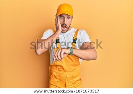 Young handsome man wearing handyman uniform over yellow background looking at the watch time worried, afraid of getting late 