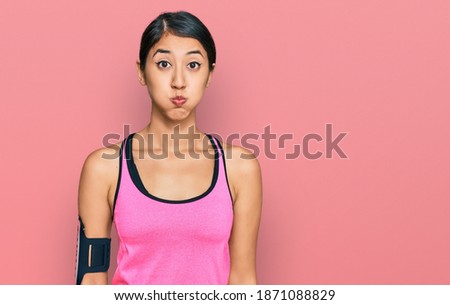 Beautiful asian young sport woman wearing sportswear and arm band puffing cheeks with funny face. mouth inflated with air, crazy expression. 