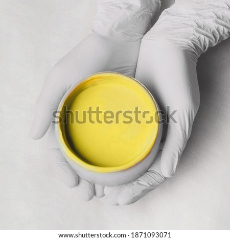 Hands in gloves hold a jar of yellow cream. Procedure in cosmetological clinic, expert with jars and bottles. Healthcare, wellness, cosmetology. Trendy color of 2021 year.