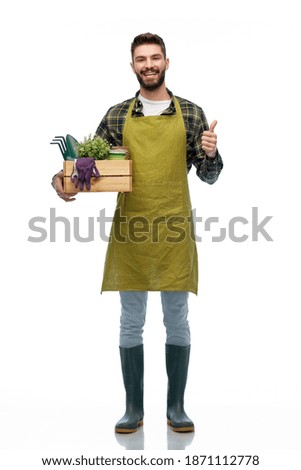 gardening, farming and people concept - happy smiling male gardener or farmer in apron and rubber boots with box of garden tools showing thumbs up over grey background