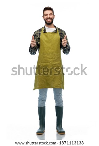 gardening, farming and people concept - happy smiling male gardener or farmer in apron and rubber boots showing thumbs up over white background