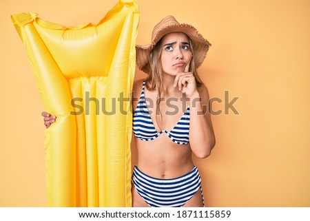 Young beautiful blonde woman wearing bikini holding floater serious face thinking about question with hand on chin, thoughtful about confusing idea 