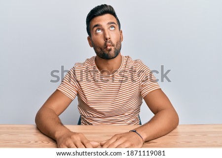 Handsome hispanic man wearing casual clothes sitting on the table making fish face with lips, crazy and comical gesture. funny expression. 