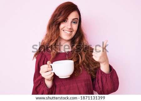 Young beautiful woman holding coffee smiling happy and positive, thumb up doing excellent and approval sign 