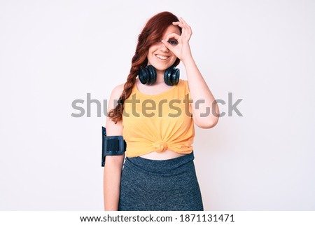 Young beautiful woman wearing gym clothes and using headphones smiling happy doing ok sign with hand on eye looking through fingers 