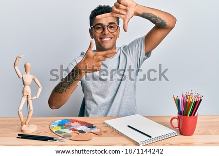 Young handsome african american man painter sitting palette and art manikin smiling making frame with hands and fingers with happy face. creativity and photography concept. 