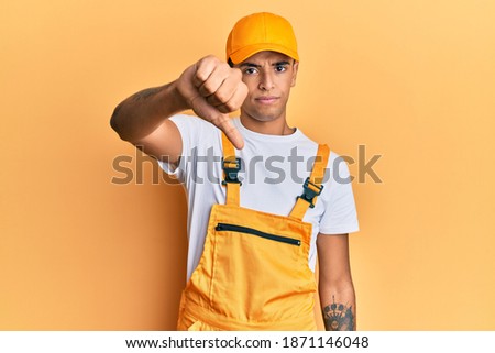 Young handsome african american man wearing handyman uniform over yellow background looking unhappy and angry showing rejection and negative with thumbs down gesture. bad expression. 
