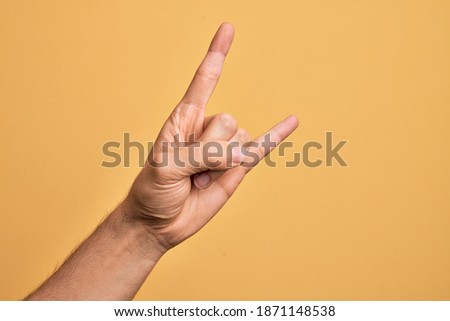 Hand of caucasian young man showing fingers over isolated yellow background gesturing rock and roll symbol, showing obscene horns gesture