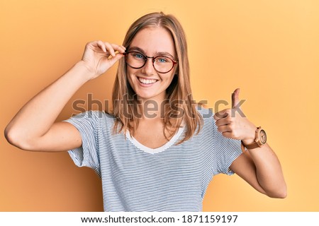 Beautiful blonde woman wearing casual clothes and holding glasses smiling happy and positive, thumb up doing excellent and approval sign 