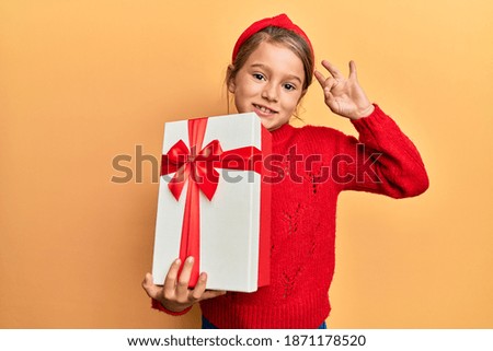 Little beautiful girl holding gift doing ok sign with fingers, smiling friendly gesturing excellent symbol 