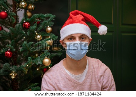 young man near a Christmas tree at home wearing a mask and hat of Santa Claus