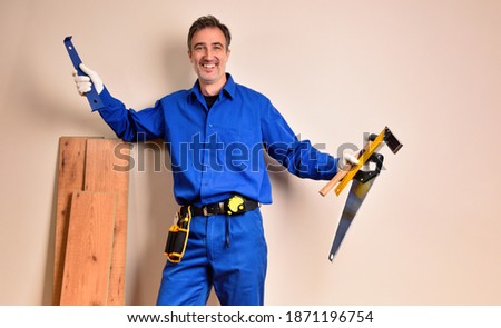 Parquet assembler in overalls smiling with tools in hands leaning on parquet slats in a room. Horizontal composition.