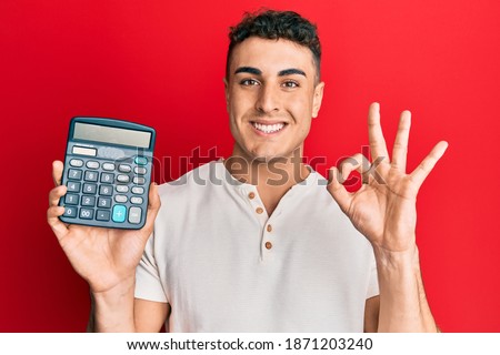 Hispanic young man showing calculator device doing ok sign with fingers, smiling friendly gesturing excellent symbol 