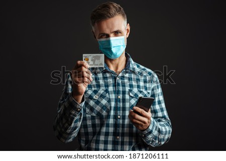 Photo of happy grey-haired man wearing face mask and using mobile phone while holding credit card isolated over grey wall background