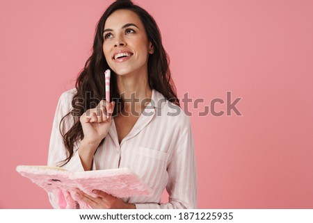 Joyful beautiful woman wearing pajama writing down notes in bunny diary isolated over pink background