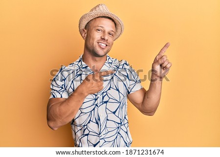 Handsome muscle man wearing summer hat smiling and looking at the camera pointing with two hands and fingers to the side. 