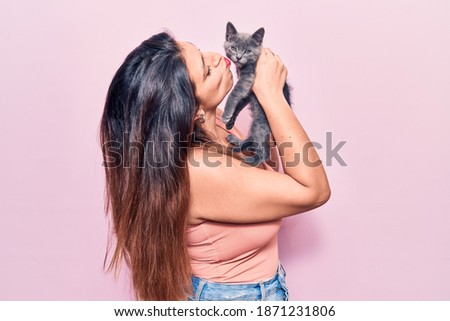 Young beautiful latin woman smilling happy. Standing with smile on face holding and kissing adorable cat over isolated pink background