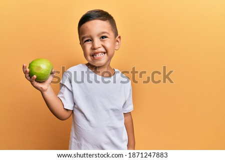 Adorable latin toddler smiling happy holding green apple looking to the camera over isolated yellow background. Royalty-Free Stock Photo #1871247883
