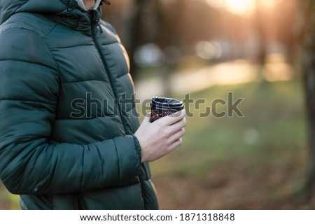 The guy holds a paper cup with coffee or tea. Have a cup of coffee. Disposable paper cup. Hot drink. Empty space. Man standing in the street holding a cup of coffee. Good morning.