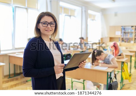 Portrait of mature woman teacher in classroom with digital tablet, smiling confident female on background of classroom with teenage students