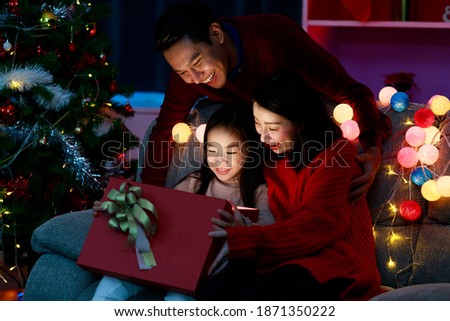 Cheerful father, mother and daughter opening and holding Christmas gift box while sitting on sofa at home. Happy Asian family on Christmas day. Merry Christmas and Happy Holidays. Christmas holiday