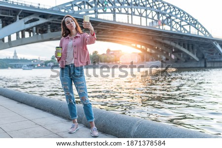 woman in summer on street, standing by river in city. Photo by smartphone, selfie and video call online. Bridge background. Jeans and pink jacket, bag. Free space for a copy of text