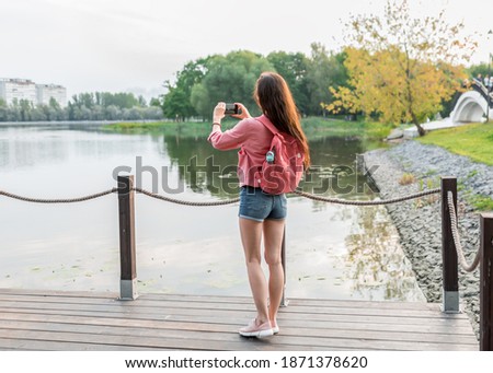 girl in a pink jacket, in summer in city, photographs lake and landscape, background of pond and river. Denim shorts on a woman. Free space for a copy of text. Green trees in autumn