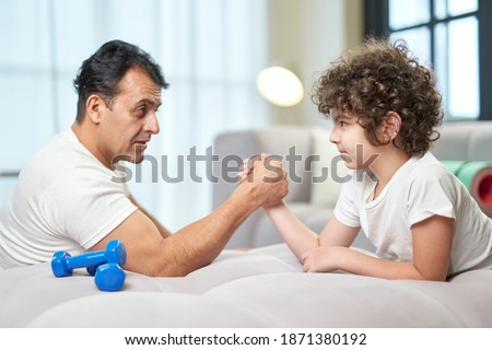 Determination. Cute latin boy and his father having fun together at home while armwrestling lying on the couch