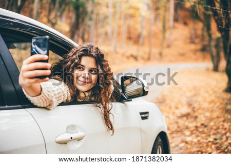 Happy curly haired young woman in white sweater makes selfie with mobile phone leaning out of modern car window in picturesque autumn forest. 