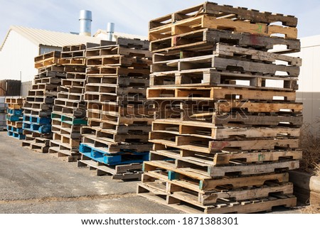 Wooden pallets stacked along a wall in a warehouse lot.  Stacks of pallets under a pale sky.  Loading dock with pallets piled high.