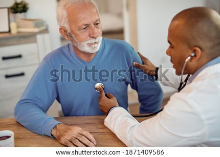 African American doctor listening senior man's heartbeat during home visit. Focus is on senior man.  Royalty-Free Stock Photo #1871409586