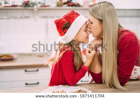 Horizontal portrait of mother and daughter. A teenage girl kisses her mother in the nose in the kitchen with gingerbread in a Christmas atmosphere in red jacket and hat. Mom and daughter relationship.