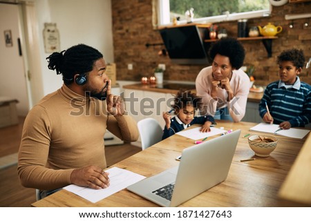 African American man silencing his wife and kids while having video call over laptop and working at home. 