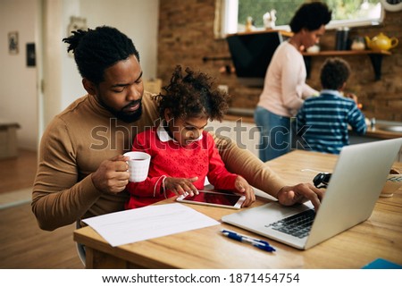 Black working father using laptop while daughter is sitting on his lap and using touchpad at home.  Royalty-Free Stock Photo #1871454754