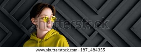 A pretty girl with brown short hair in yellow round sunglasses stands and looks away. She is wearing a yellow illuminating hoody. Background is ultimate gray relief geometric striped wall.