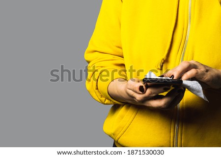 Hands wipe the screen of smartphone with disinfectant cloth. Woman is wearing yellow hoodie on gray background, trendy colors of 2021