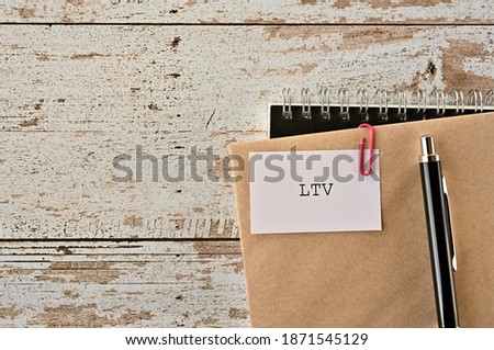 There is a piece of paper with "LTV" written on it. It was an abbreviation for Life Time Value. It was on top of white damaged wood table with a envelope and a notebook.