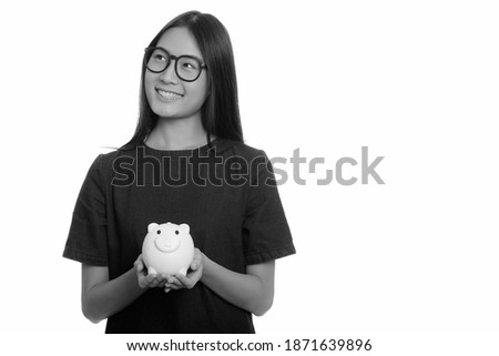 Young happy Asian teenage girl smiling and holding piggy bank while thinking