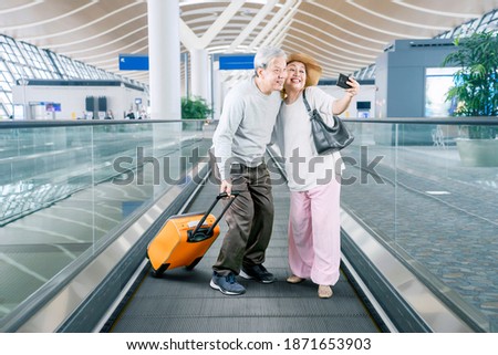 Senior couple taking a selfie photo together by using a smartphone while holding a luggage on the escalator of airport terminal