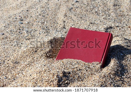 Red book on the sand on a blurry background, covered with sand, buried in the sand