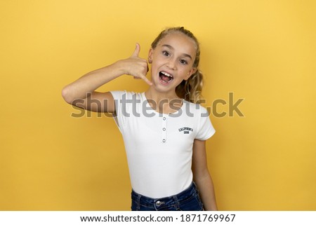 Young beautiful child girl standing over isolated yellow background smiling doing phone gesture with hand and fingers like talking on the telephone