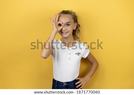 Young beautiful child girl standing over isolated yellow background doing ok gesture shocked with smiling face, eye looking through fingers